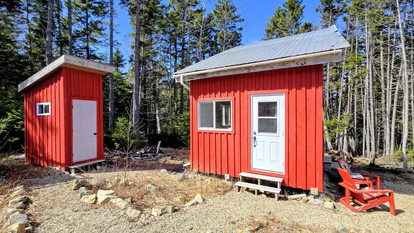 Two small, red, wooden cabins with white doors and metal roofs sit in a forest clearing on a sunny day. A red Adirondack chair sits near the cabin on the right.
