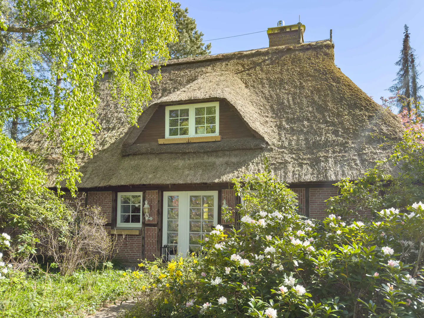 Exterior view of a brick house with a thatched roof, white windows and door, surrounded by lush greenery and flowers.