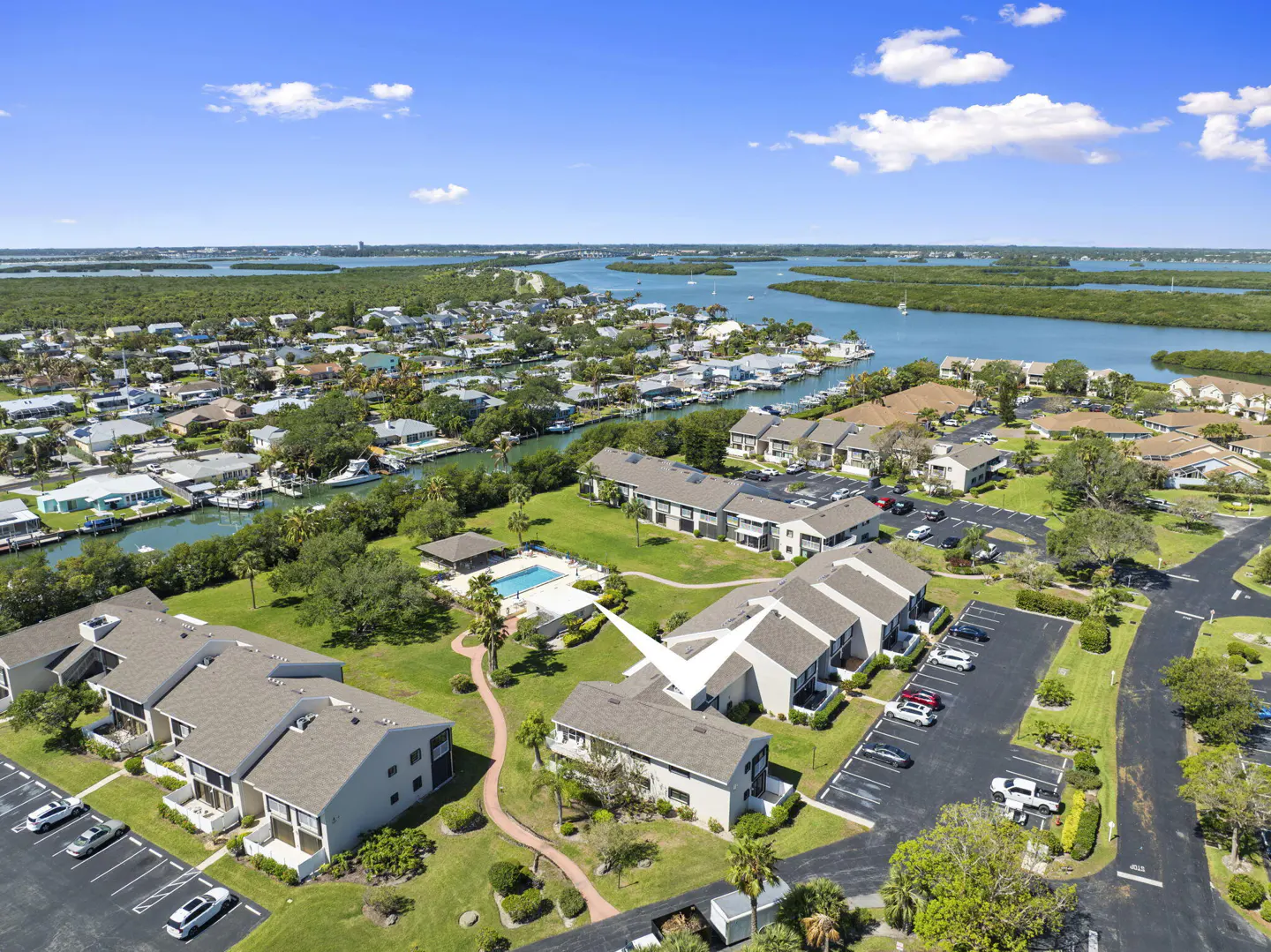 Aerial view of condos with a pool, parking lot, and waterfront access under a blue sky.