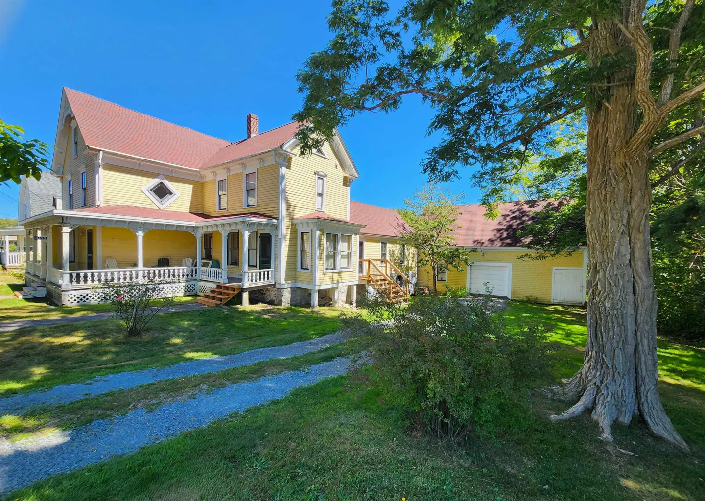 A yellow, two-story house with a red roof and white porch, surrounded by green grass and trees under a blue sky.