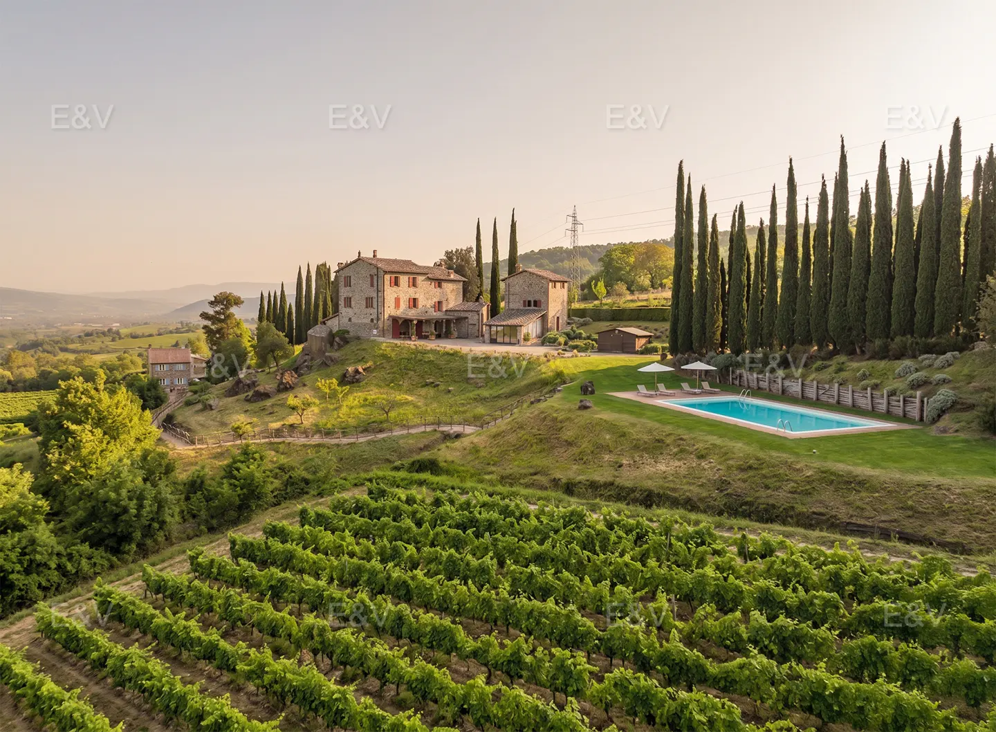 Tuscan villa with a pool and vineyard. Stone house with red shutters sits on a hill, surrounded by tall cypress trees and green vines.