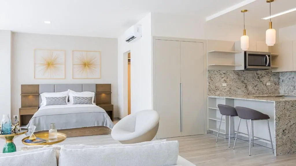 Bright studio apartment with a bed, white sofa, and kitchen with granite countertops and two gray bar stools.