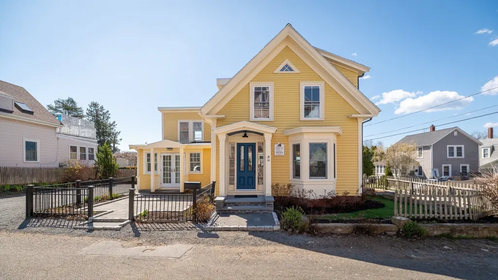 A yellow two-story house with a blue door and white trim on a sunny day. Black metal fence in front.