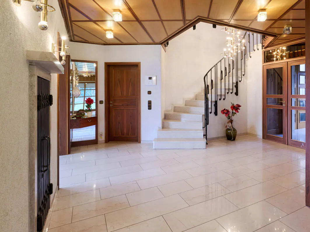 Elegant foyer with marble floors, a wooden ceiling, and a winding staircase with a chandelier. A vase of red flowers sits at the base of the stairs.