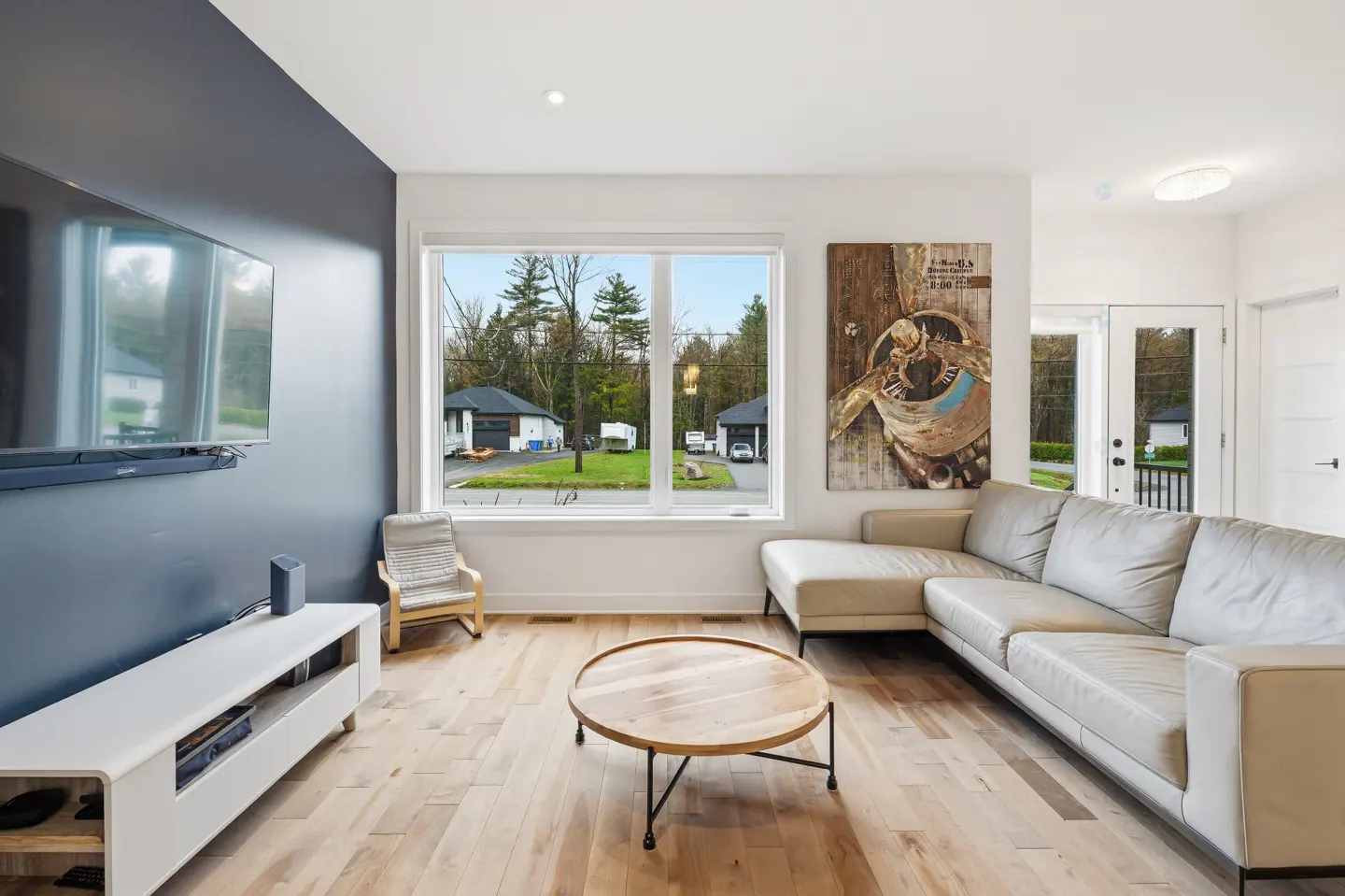 Living room with hardwood floors, a beige sectional sofa, and a round wooden coffee table. A large window overlooks a green lawn and houses.