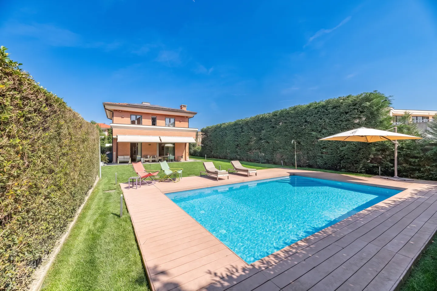 A backyard with a pool, wooden deck, lounge chairs, and a two-story house with an awning.