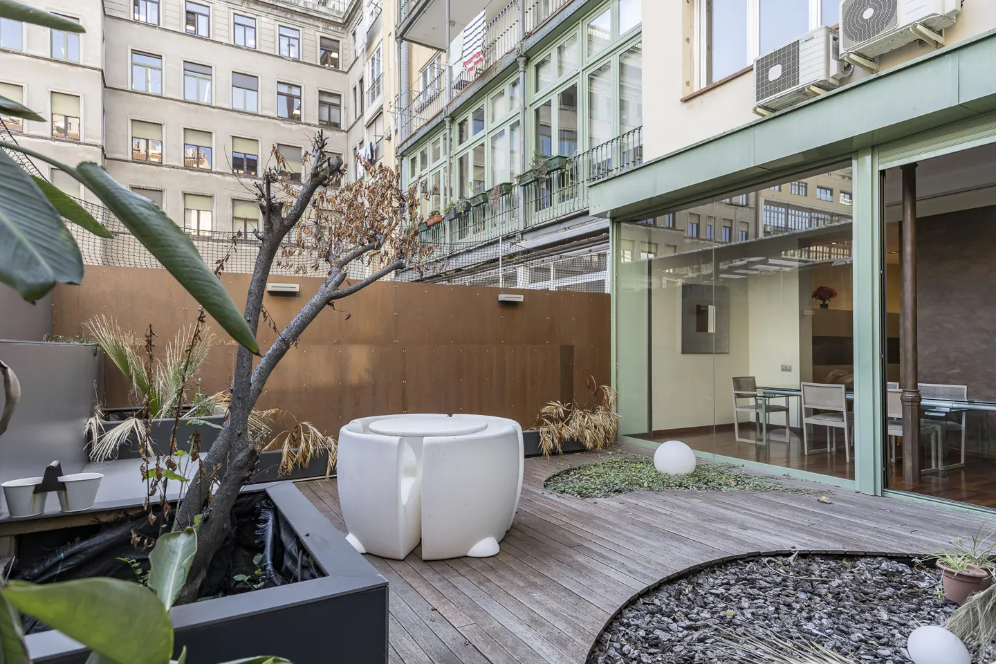 Modern urban terrace with wooden decking, white sculptural seating, a small tree, and glass walls looking into a dining room.