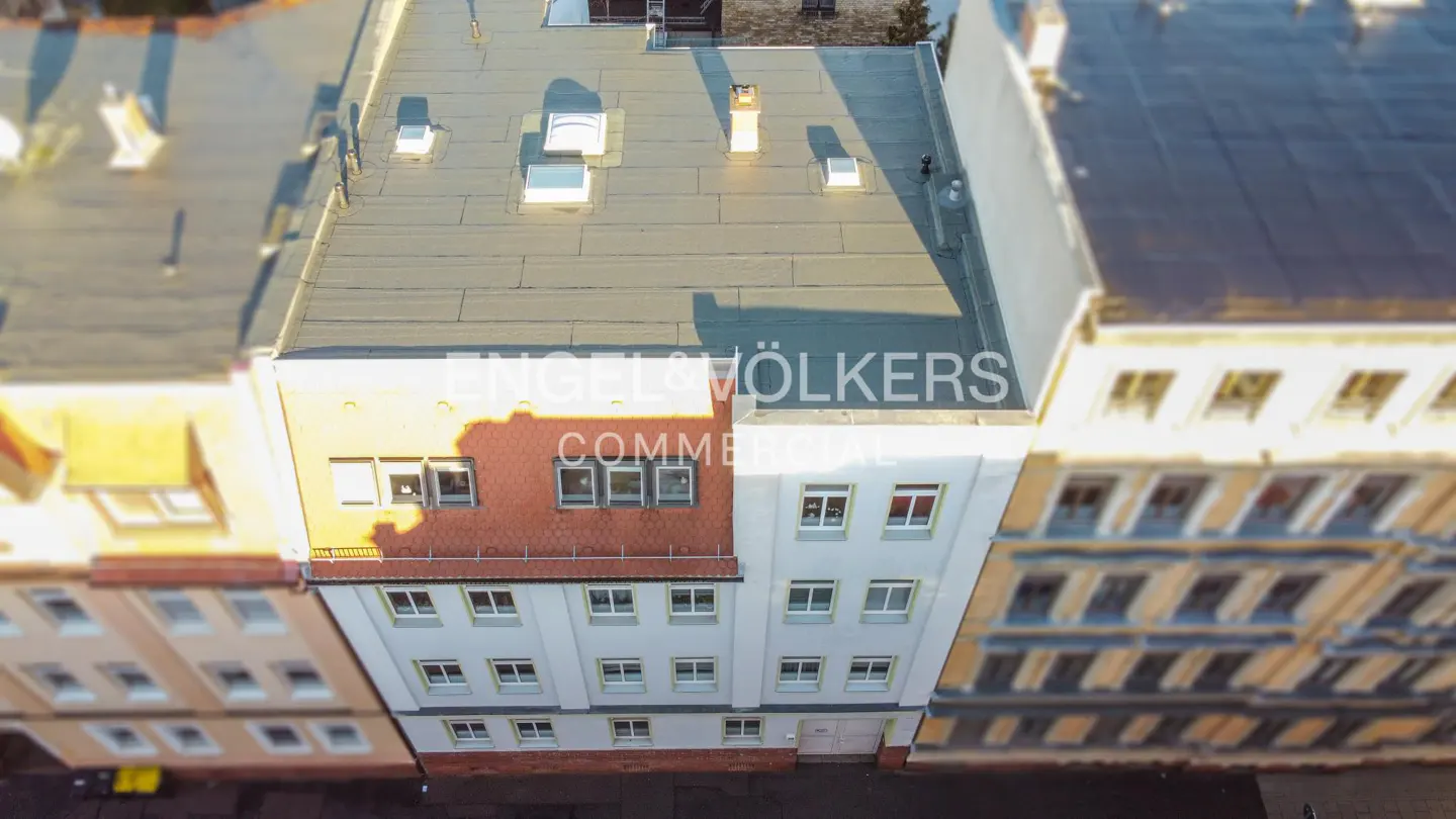Aerial view of a white building with many windows and a gray roof, next to other buildings.
