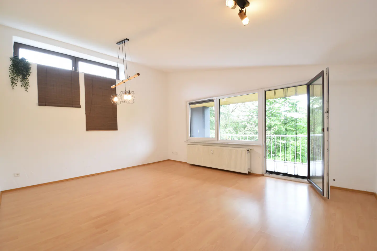 Bright, empty room with wood floors, white walls, and large windows. A modern light fixture hangs near the window with brown blinds. Balcony door is open.
