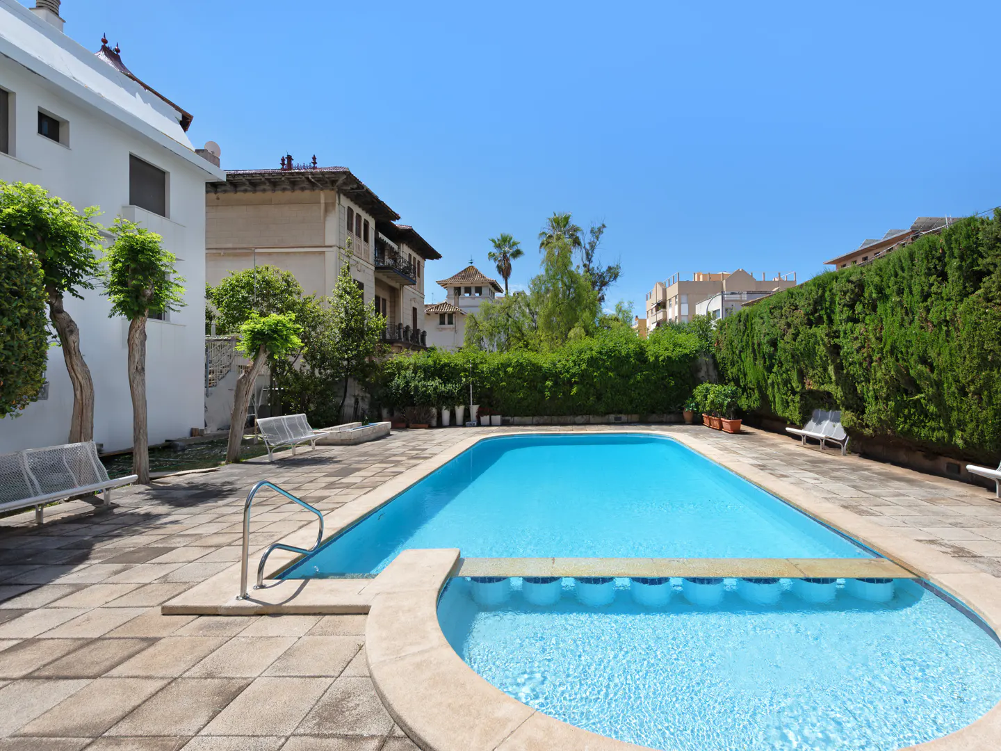 Outdoor pool with clear blue water, surrounded by stone patio, benches, and lush green hedges. Buildings visible in the background.