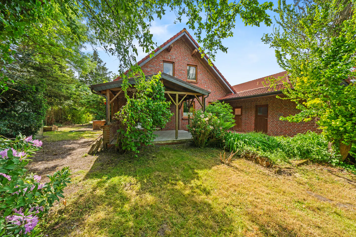 Red brick house with a wooden patio, surrounded by lush green trees and a grassy yard under a bright blue sky.