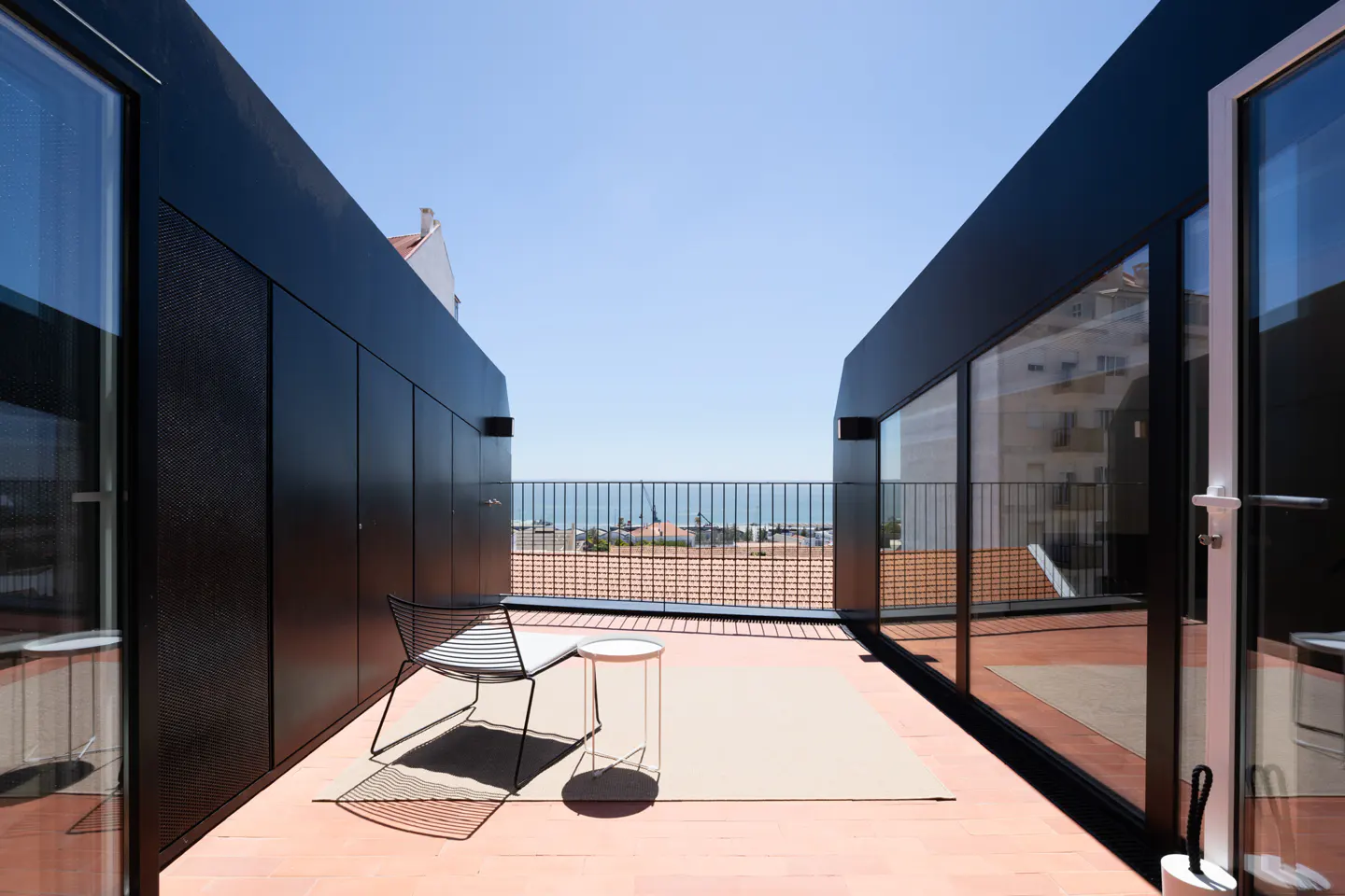 Modern rooftop terrace with black walls, a wire chair, small table, and a metal railing overlooking a coastal city view.