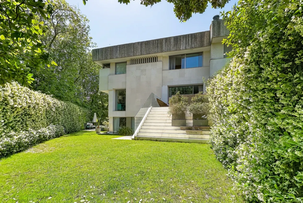 Modern two-story house with a concrete facade, large windows, and a staircase leading to the entrance, surrounded by green lawn and hedges.