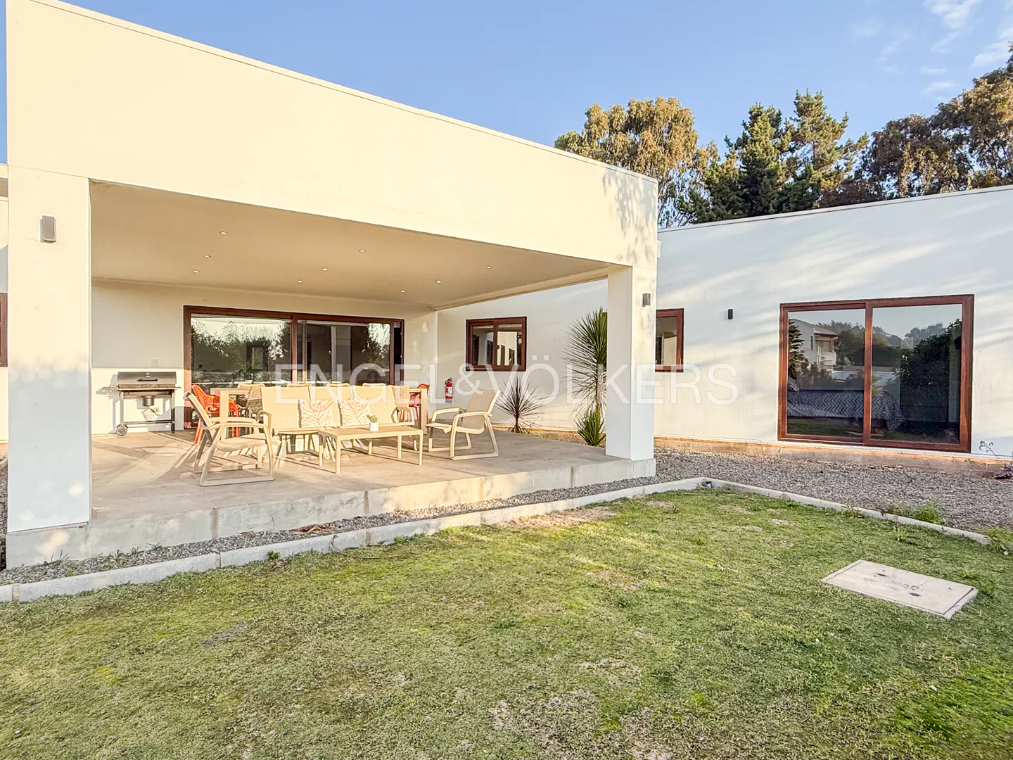 Modern white house with a covered patio, outdoor seating, and a grassy backyard under a clear blue sky.