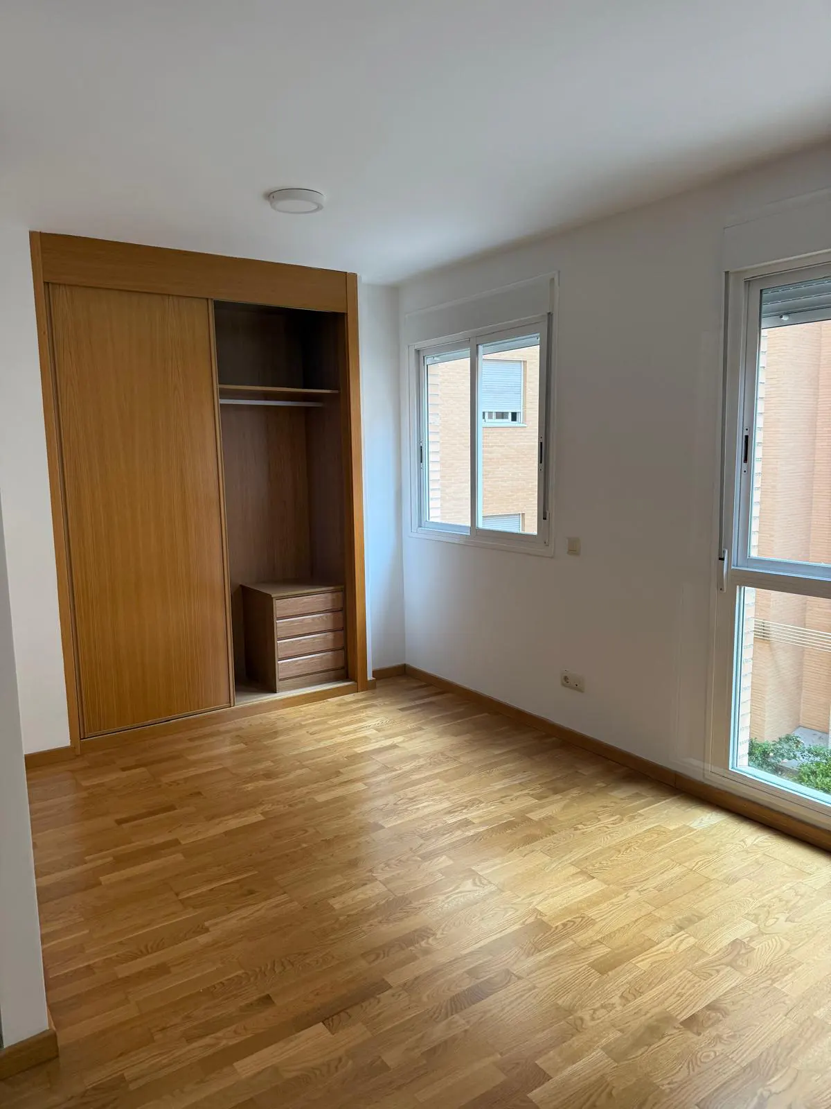 Bright, empty bedroom with wood floors, white walls, and two windows. A large, built-in wood closet with sliding doors is on the left.
