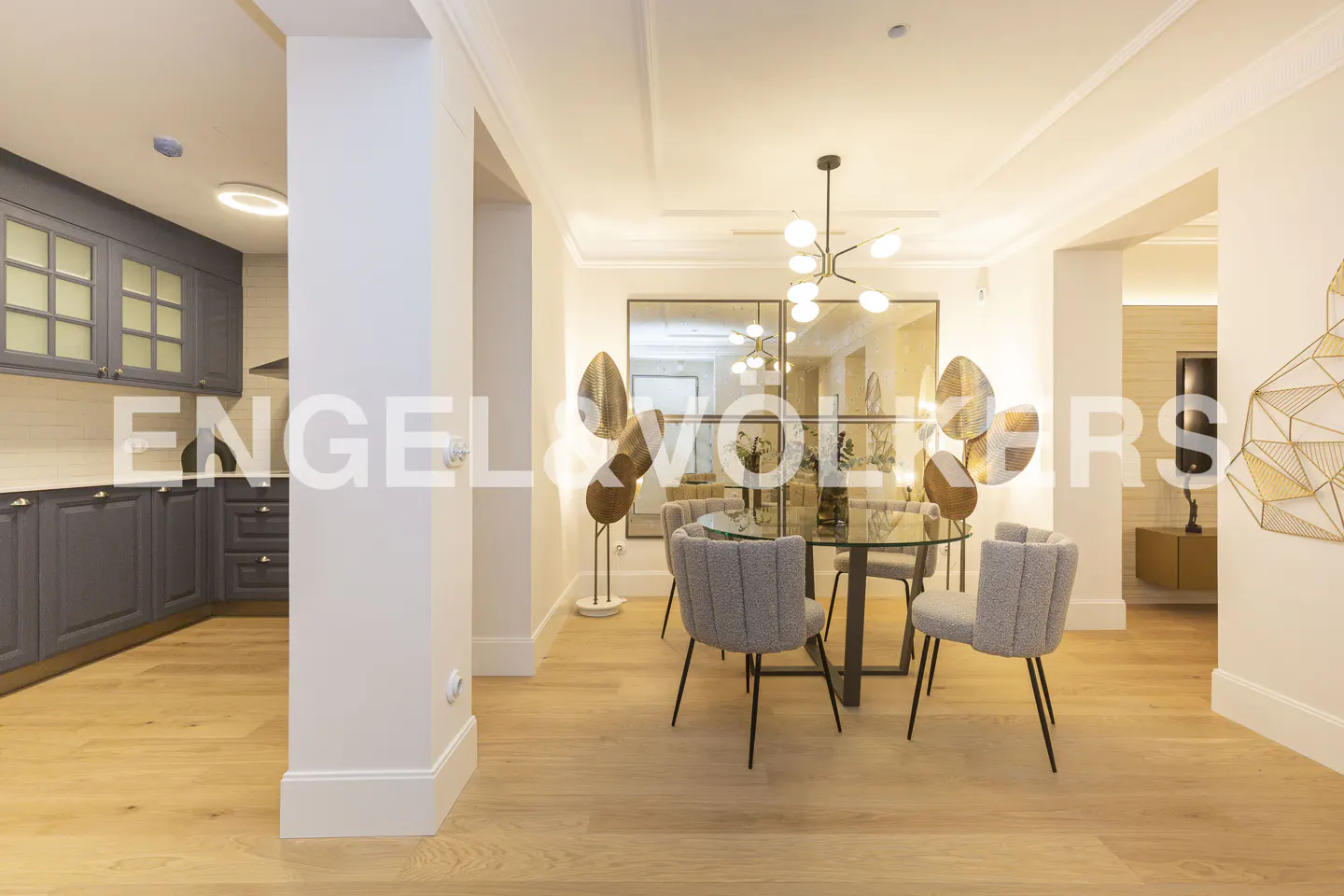 Bright, open-concept kitchen and dining area with light wood floors. Gray cabinets, glass table, and modern light fixture.