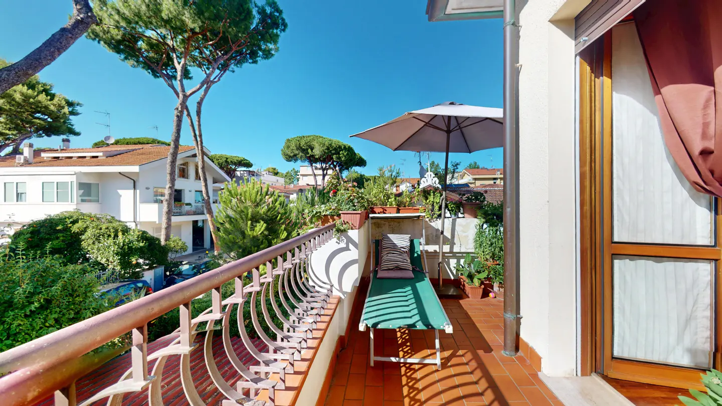 Balcony with a green lounge chair, umbrella, and plants overlooking a neighborhood with trees under a blue sky.