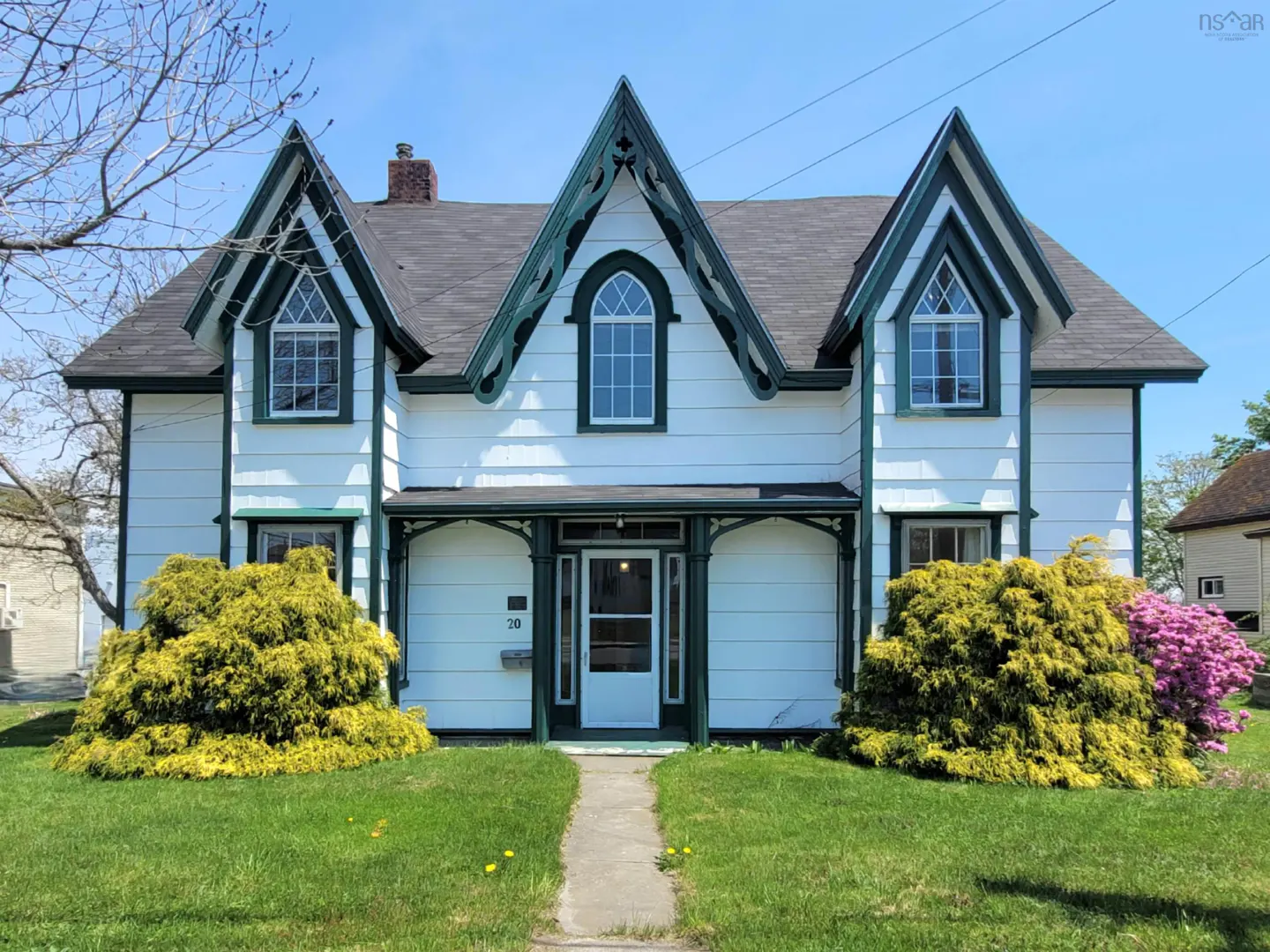 Two-story white house with green trim, black roof, and peaked gables. A stone path leads to the front door. Bushes flank the entrance.