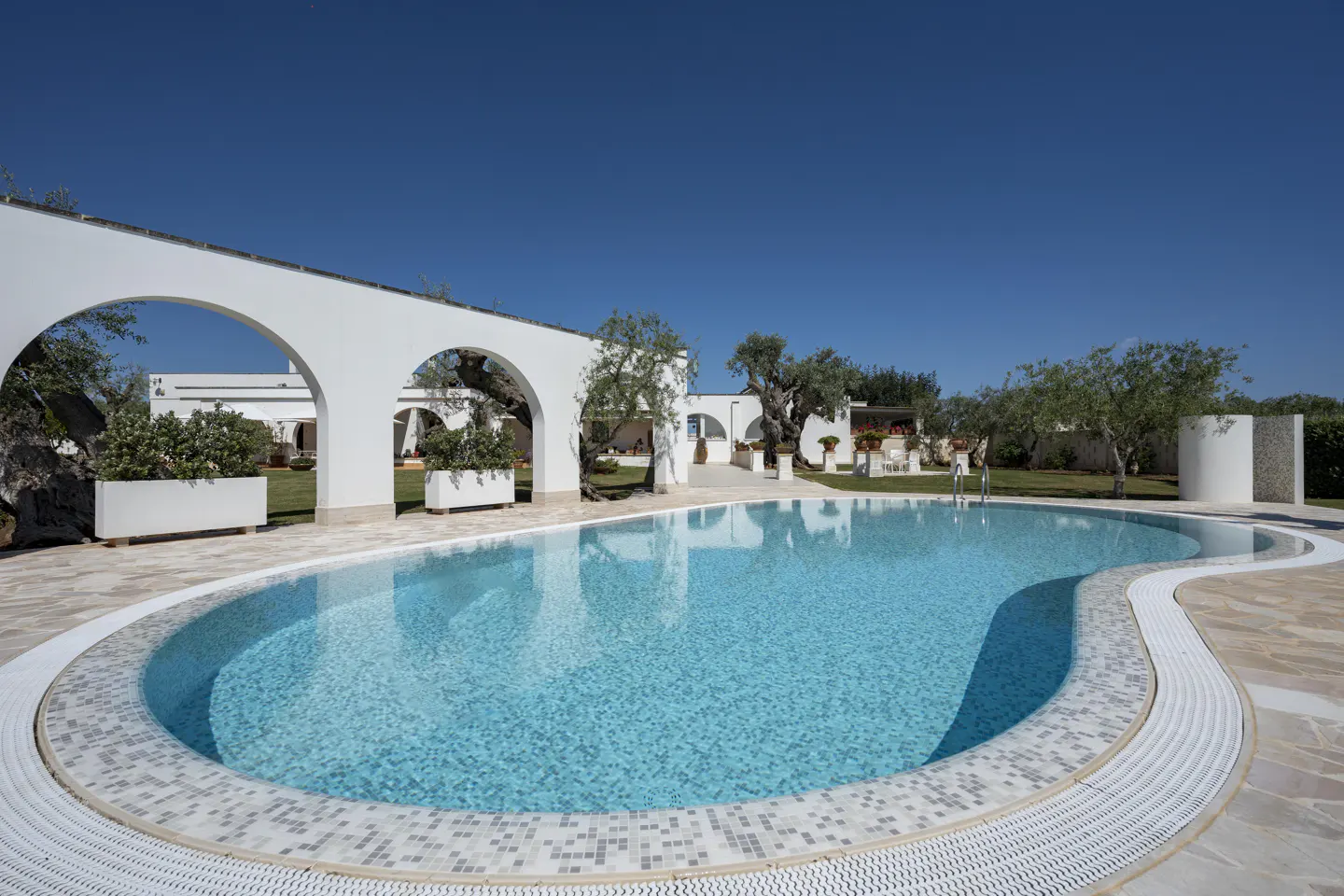 A turquoise pool with white arches and planters under a clear blue sky.