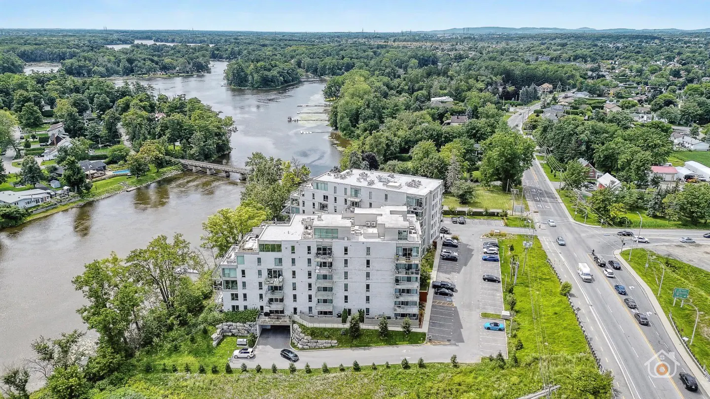 Aerial view of a modern white condo building next to a river, with a parking lot and road nearby.
