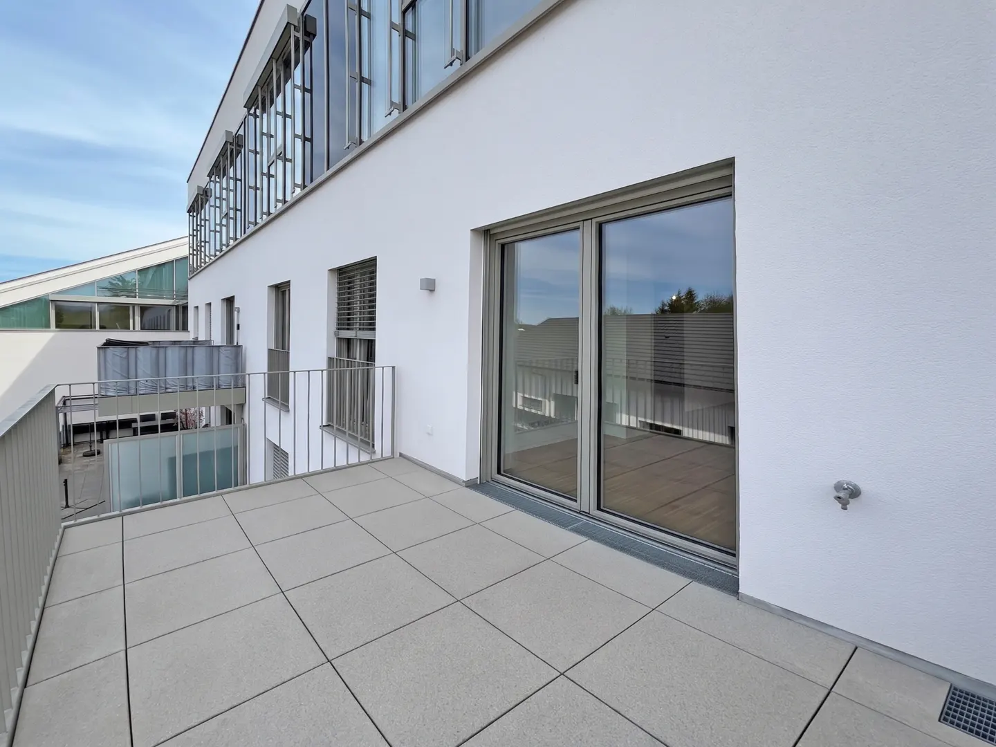 A modern apartment balcony with gray tiled floor, metal railing, and sliding glass doors on a white building.