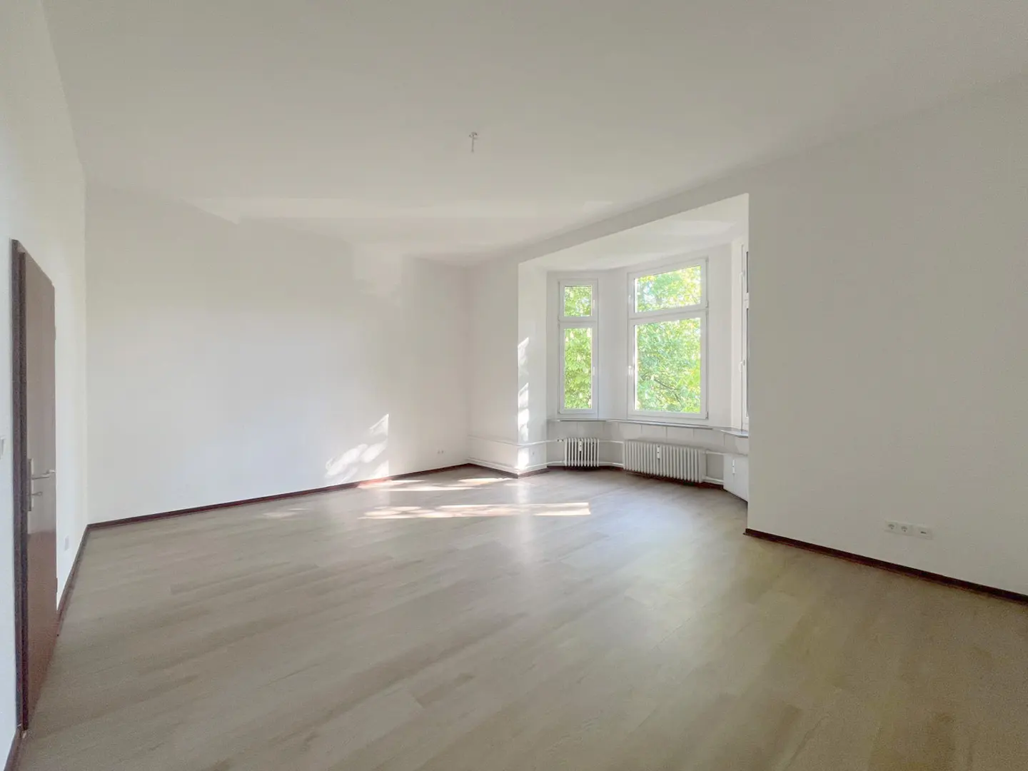 Bright, empty room with white walls, light wood floors, and a bay window showing green trees outside. A radiator sits beneath the window.