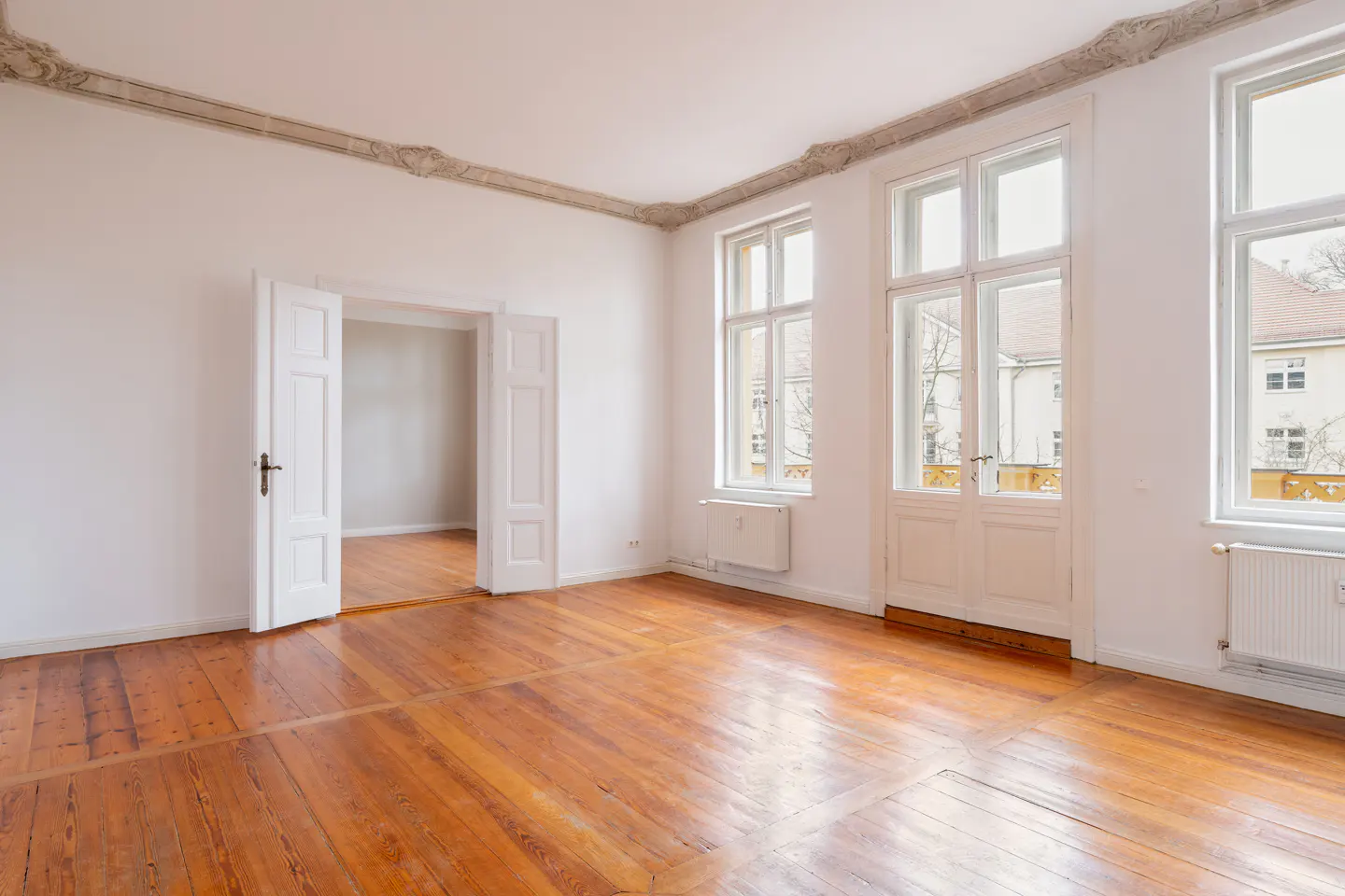 Bright, empty room with hardwood floors, white walls, and ornate ceiling trim. Open doorway leads to another room. Windows let in natural light.