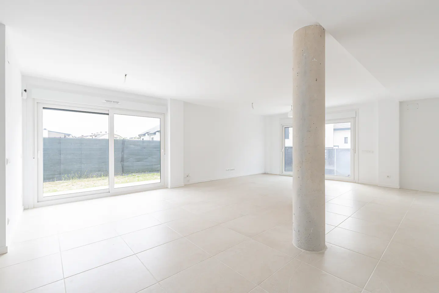 Bright, empty room with white walls, tile floor, and a concrete support column. Large sliding glass doors offer a view of the yard.