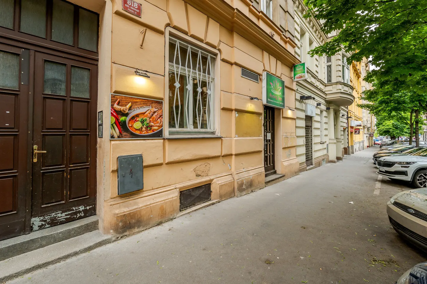 Street view of a tan building with a brown door, a window with bars, and a Thai restaurant sign. Cars are parked along the street.