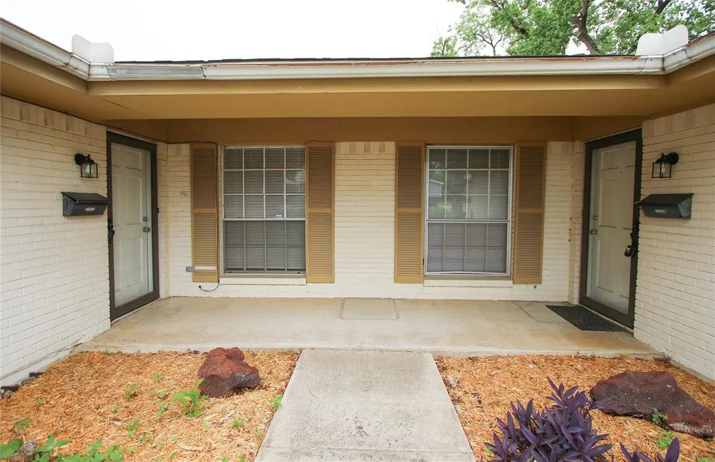 Exterior view of a duplex with white brick, tan trim, two white doors, and two windows with tan shutters. A concrete walkway leads to the entrance.
