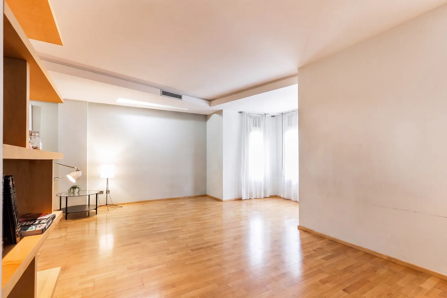 Bright, empty living room with hardwood floors, white walls, and sheer curtains. A glass table and lamp sit near a built-in wooden shelf.