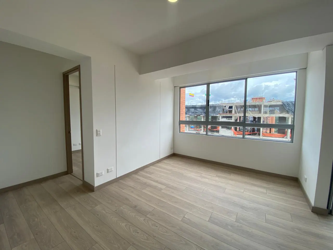 A bright, empty room with light wood floors and white walls. A large window shows a building under construction.