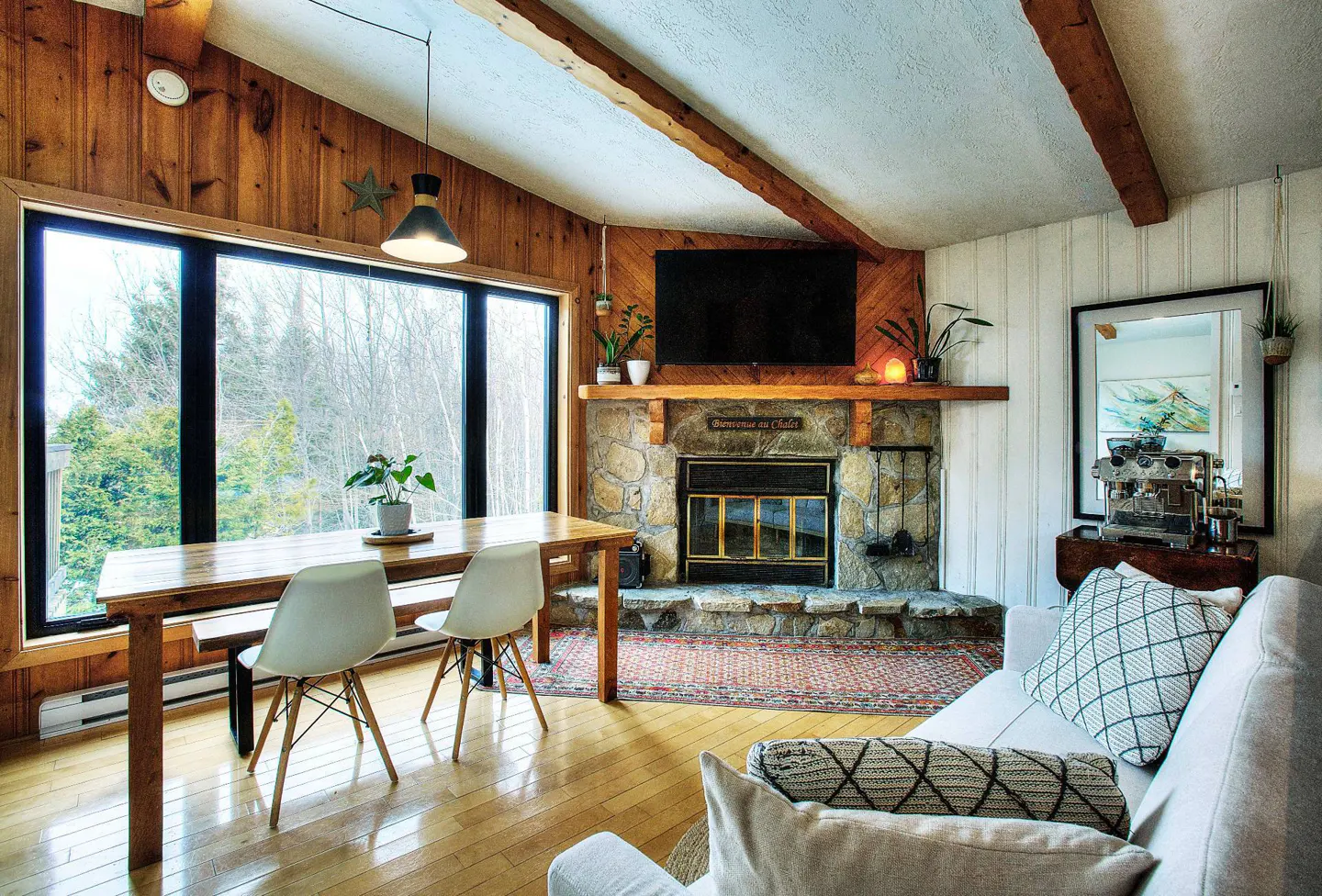Living room with wood walls, stone fireplace, and large window overlooking trees. A wooden table and white chairs sit near the window.