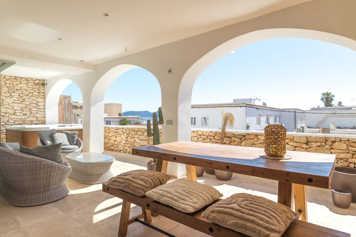 Outdoor patio with white arches framing a sea view. Wicker furniture, a wooden table with cushions, and stone walls create a relaxed atmosphere.