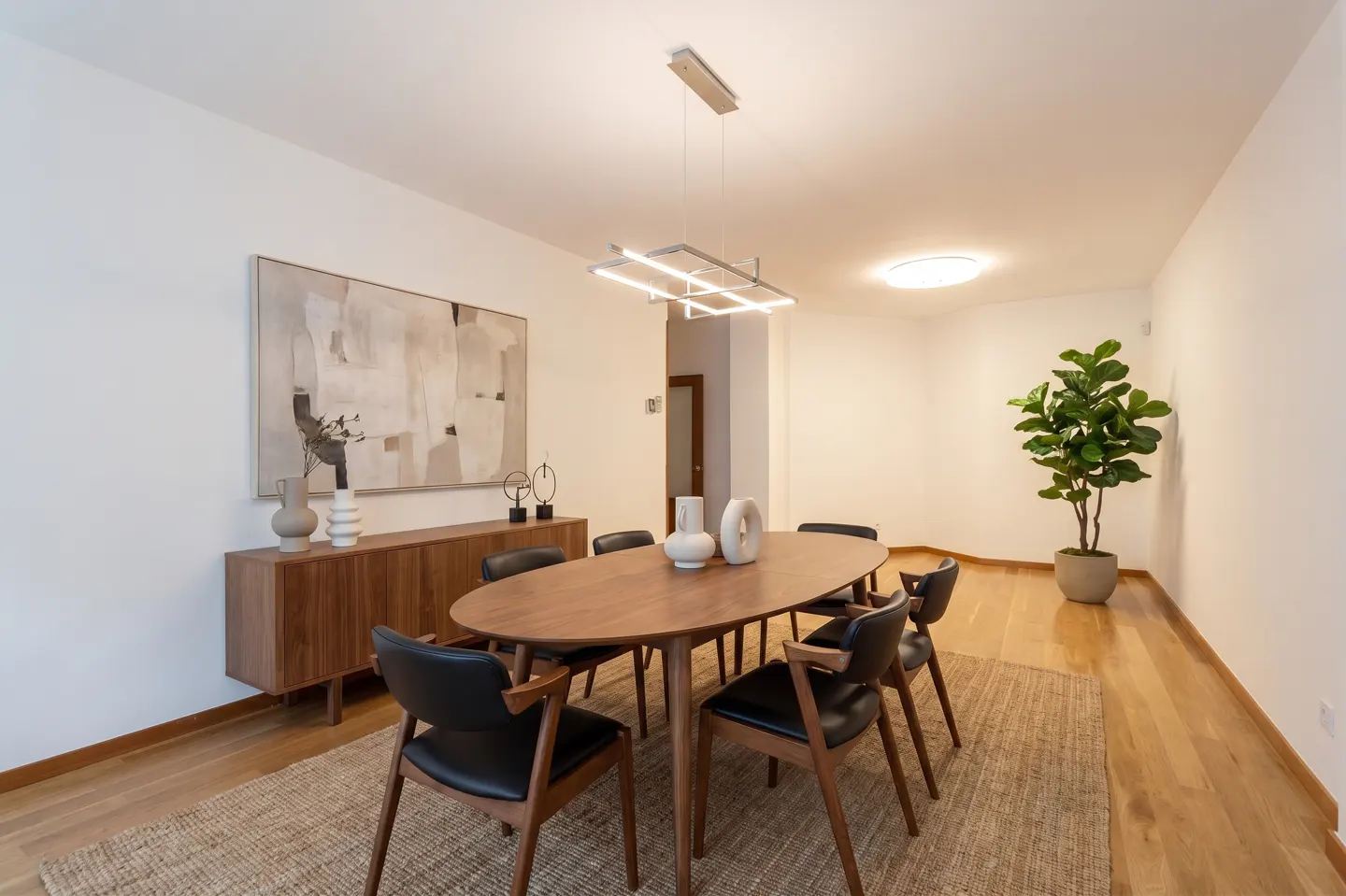 Bright dining room with wood floors, oval table, black chairs, and a modern light fixture. A large painting hangs above a wood cabinet.