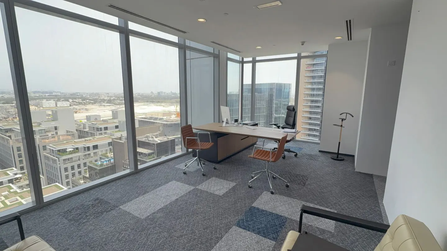 Bright office space with floor-to-ceiling windows overlooking a city. A desk with chairs faces the view. Gray and blue carpet tiles cover the floor.