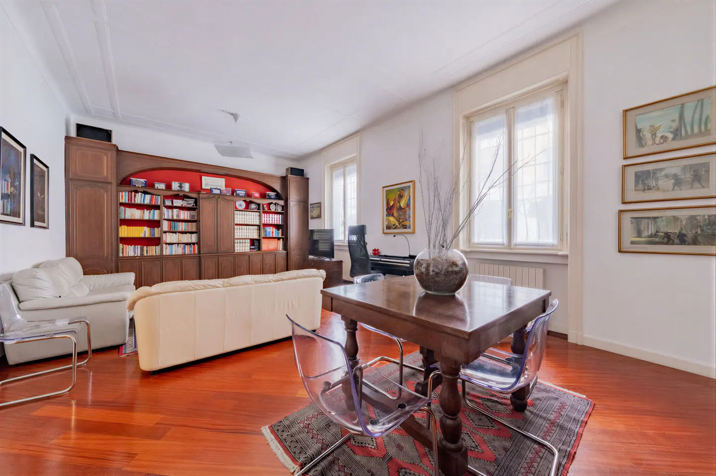 Living room with hardwood floors, a cream sofa, a wooden dining table with clear chairs, and a large built-in bookshelf.