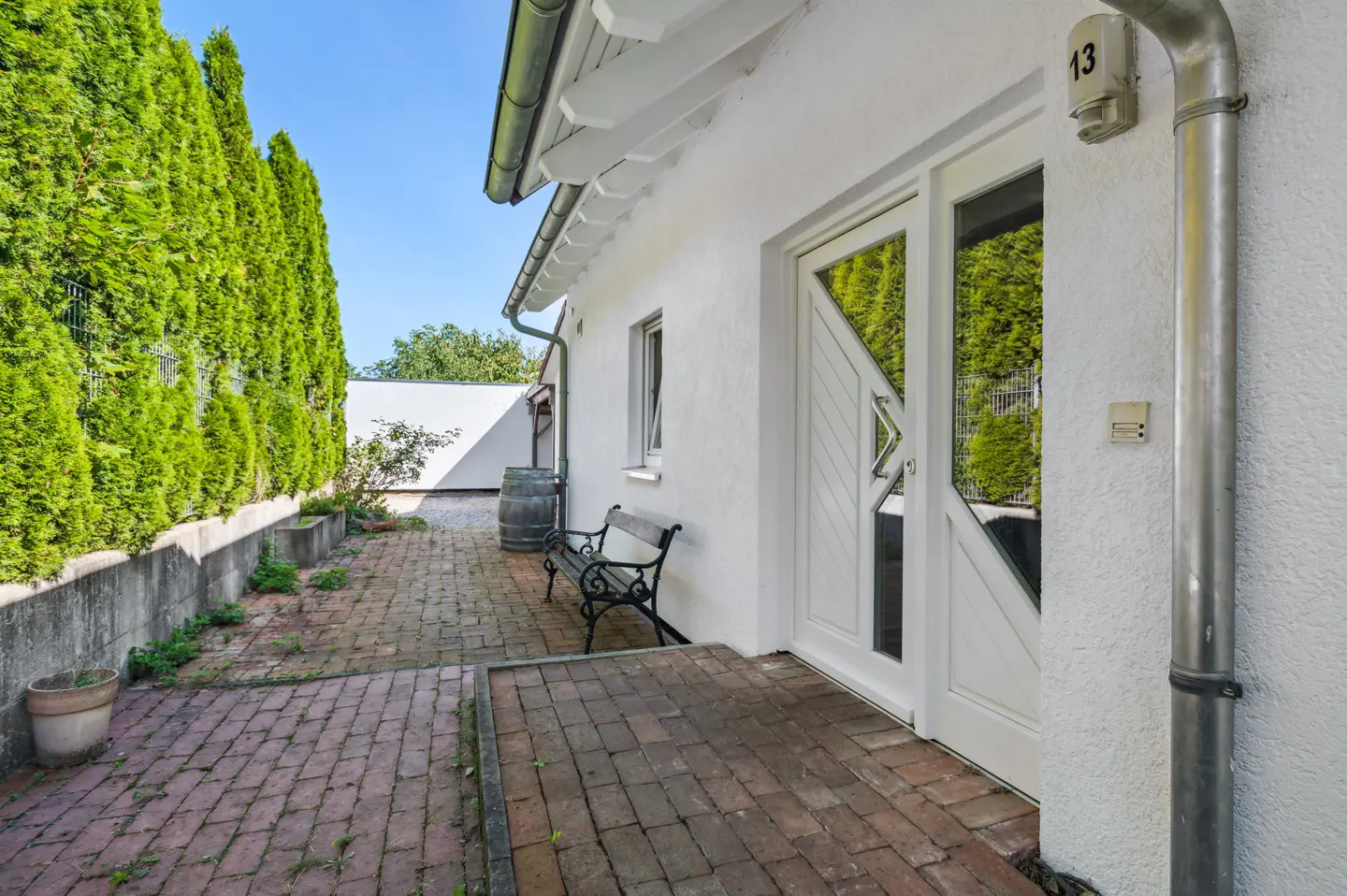 Exterior view of a white house with a brick patio, bench, and tall green hedges. The front door is white with glass panels. A rain gutter runs down the side of the house.