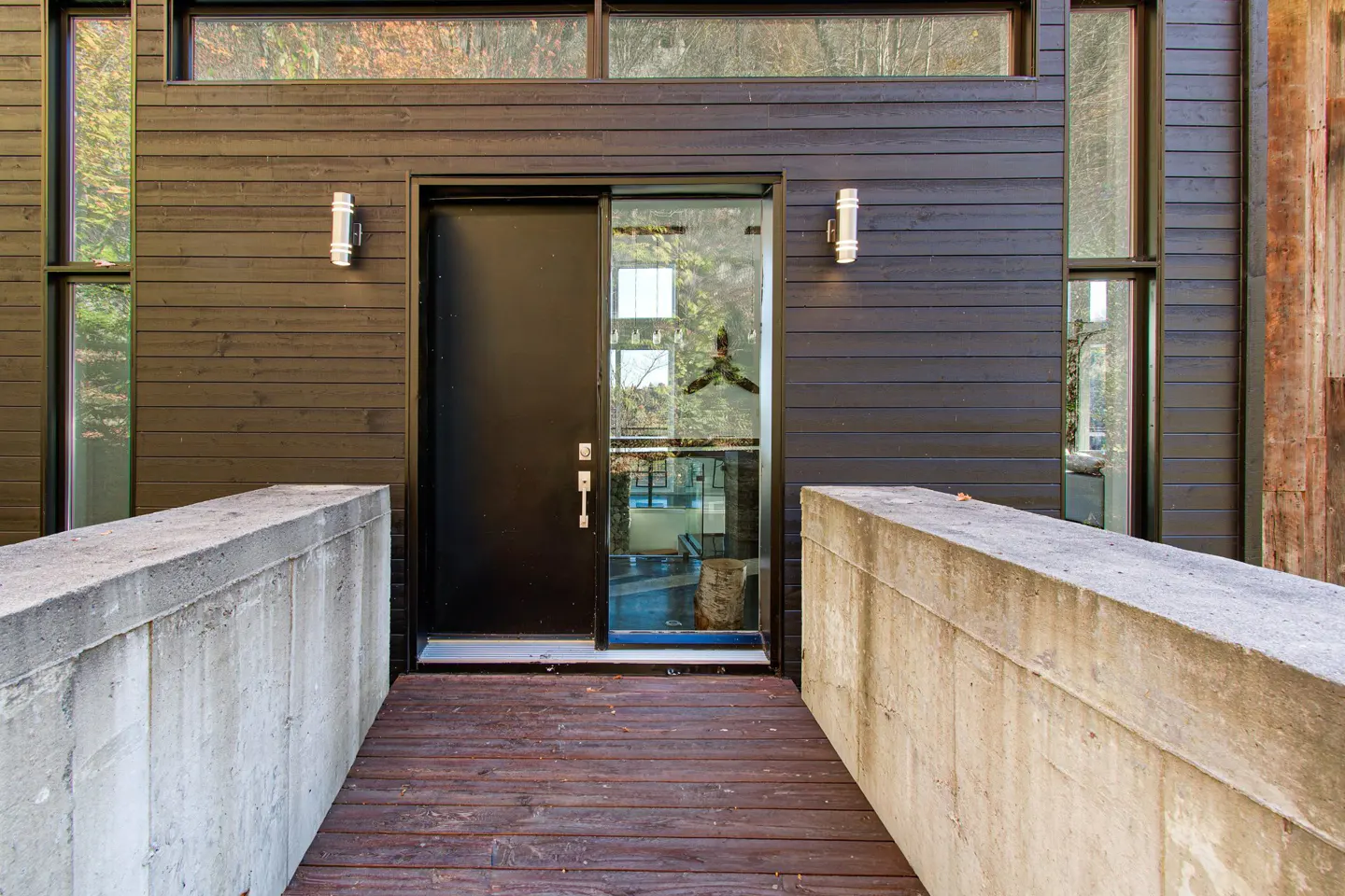 Modern home exterior with a black door, glass panel, and dark wood siding. Concrete retaining walls flank a wooden walkway.