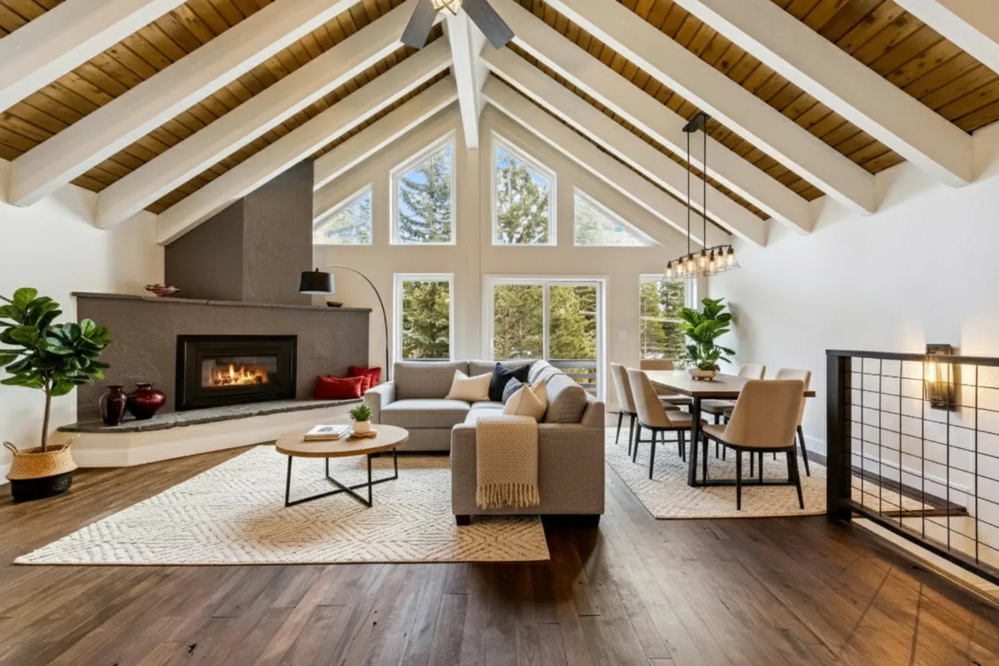 Open-concept living room with vaulted wood-beam ceiling, fireplace, grey sectional sofa, and dining area with large windows.