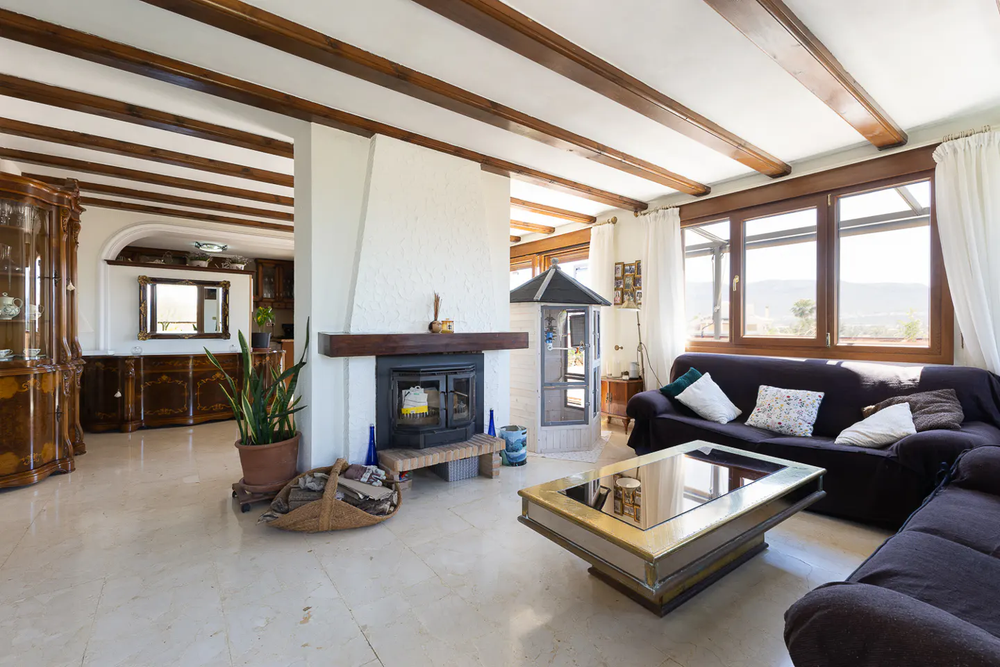 Bright living room with a fireplace, dark brown sofa, and a glass coffee table. Large windows offer a view of the outside.