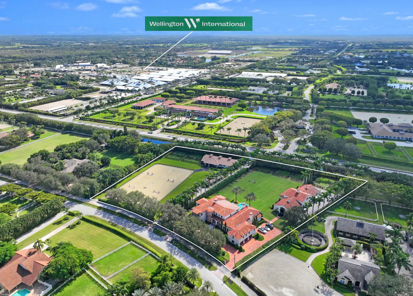 Aerial view of a large estate in Wellington, Florida, with a red-tiled roofed mansion, horse paddocks, and lush green landscaping.
