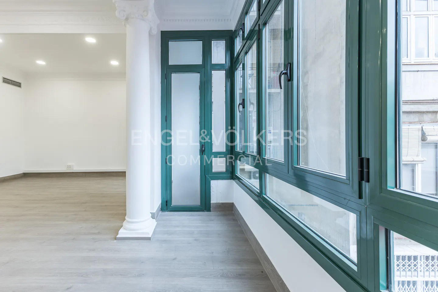 Bright room with white walls, light wood floors, and a green-framed window and door leading to a balcony. A white column stands near the window.