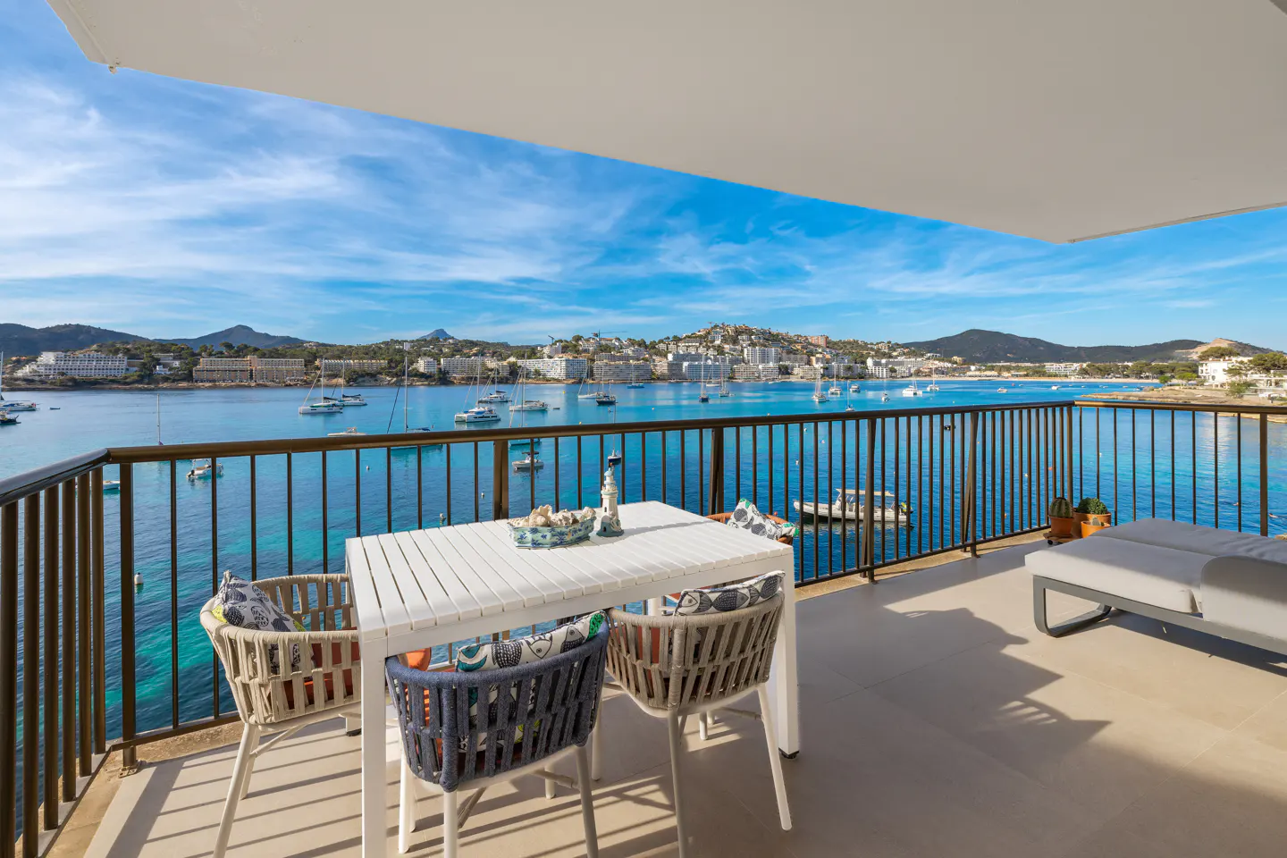 Balcony view of a blue bay. A white table and chairs sit on the patio, with a lounge chair to the right. Boats dot the water.