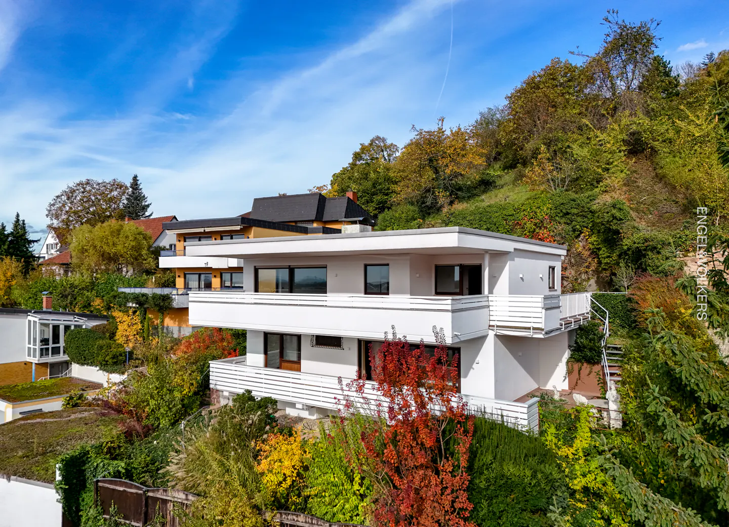 Two-story white house with balconies, surrounded by lush greenery on a hillside under a blue sky.