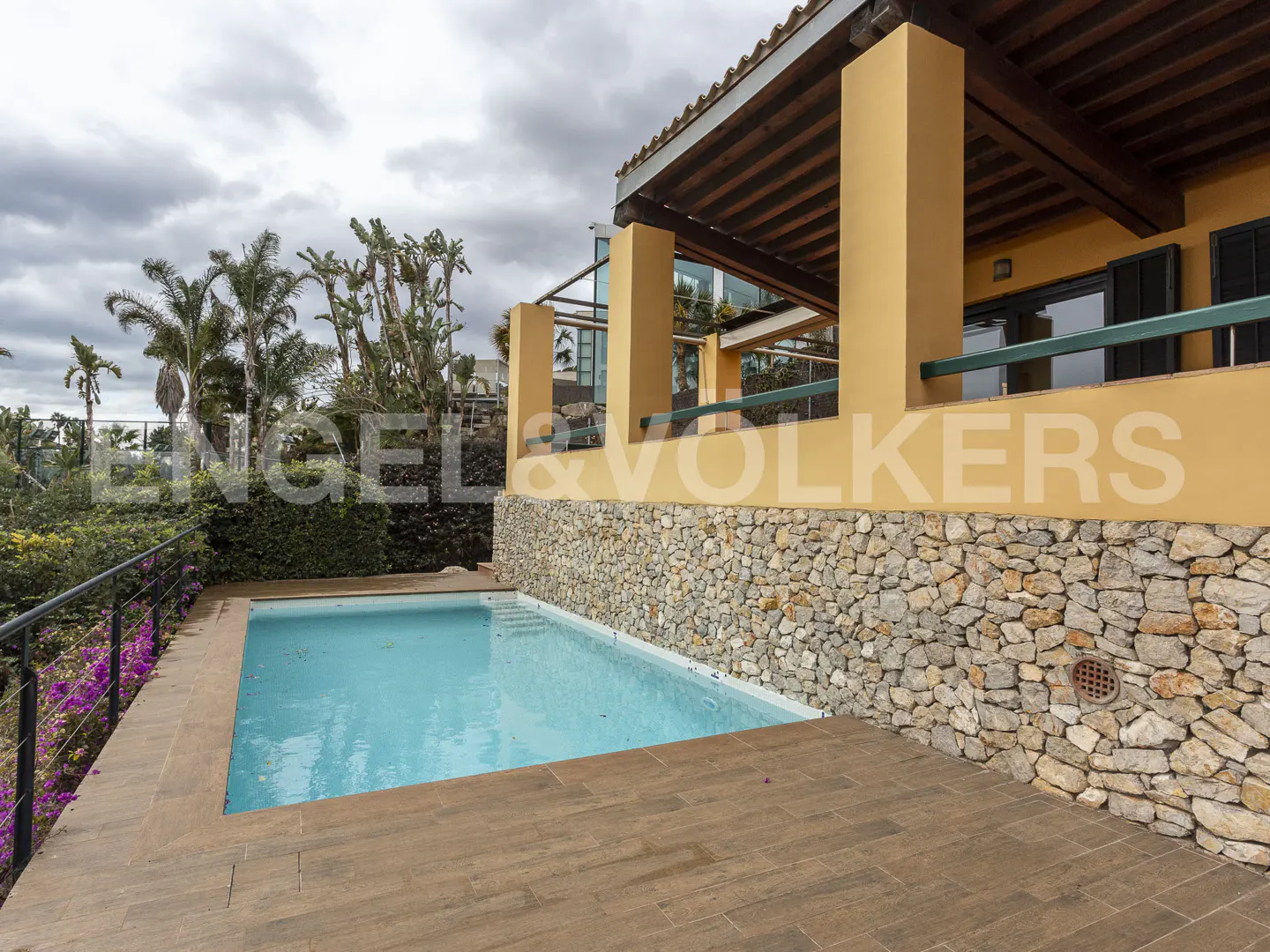 A rectangular swimming pool with blue water sits next to a stone-walled patio and a yellow house with a wooden pergola.