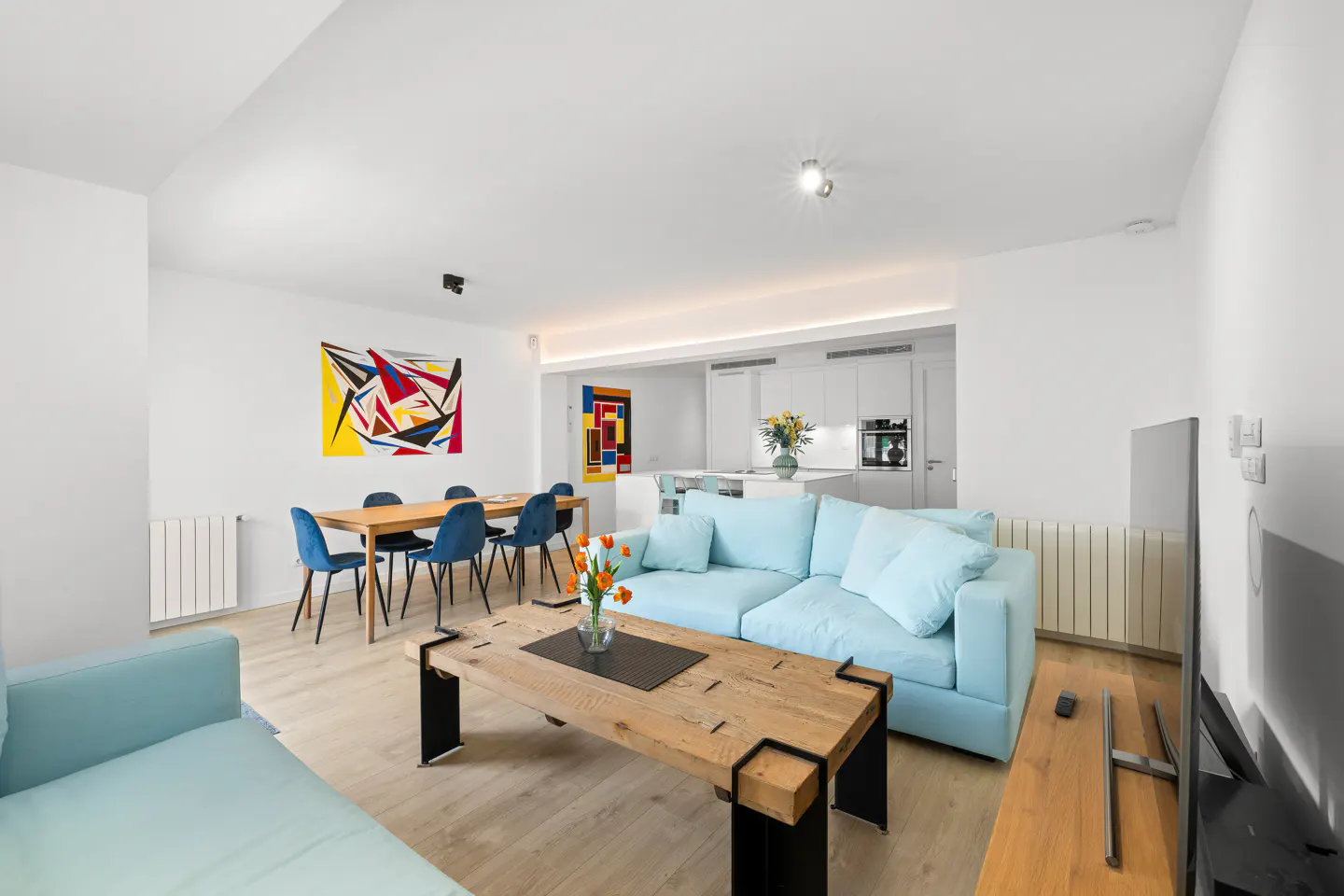 Bright, modern living room with light blue sofas, wood table, dining set, and abstract art. White walls and open kitchen.