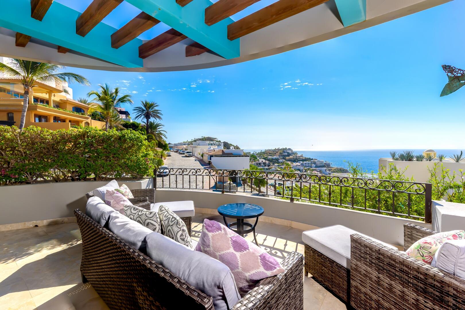 Wicker patio furniture on a balcony with a turquoise pergola, overlooking a coastal town and the ocean under a clear sky. [AI-generated text]