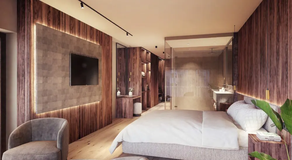 Modern bedroom with wood paneling, a gray chair, and a bed with white linens. A glass-enclosed bathroom is visible in the background.