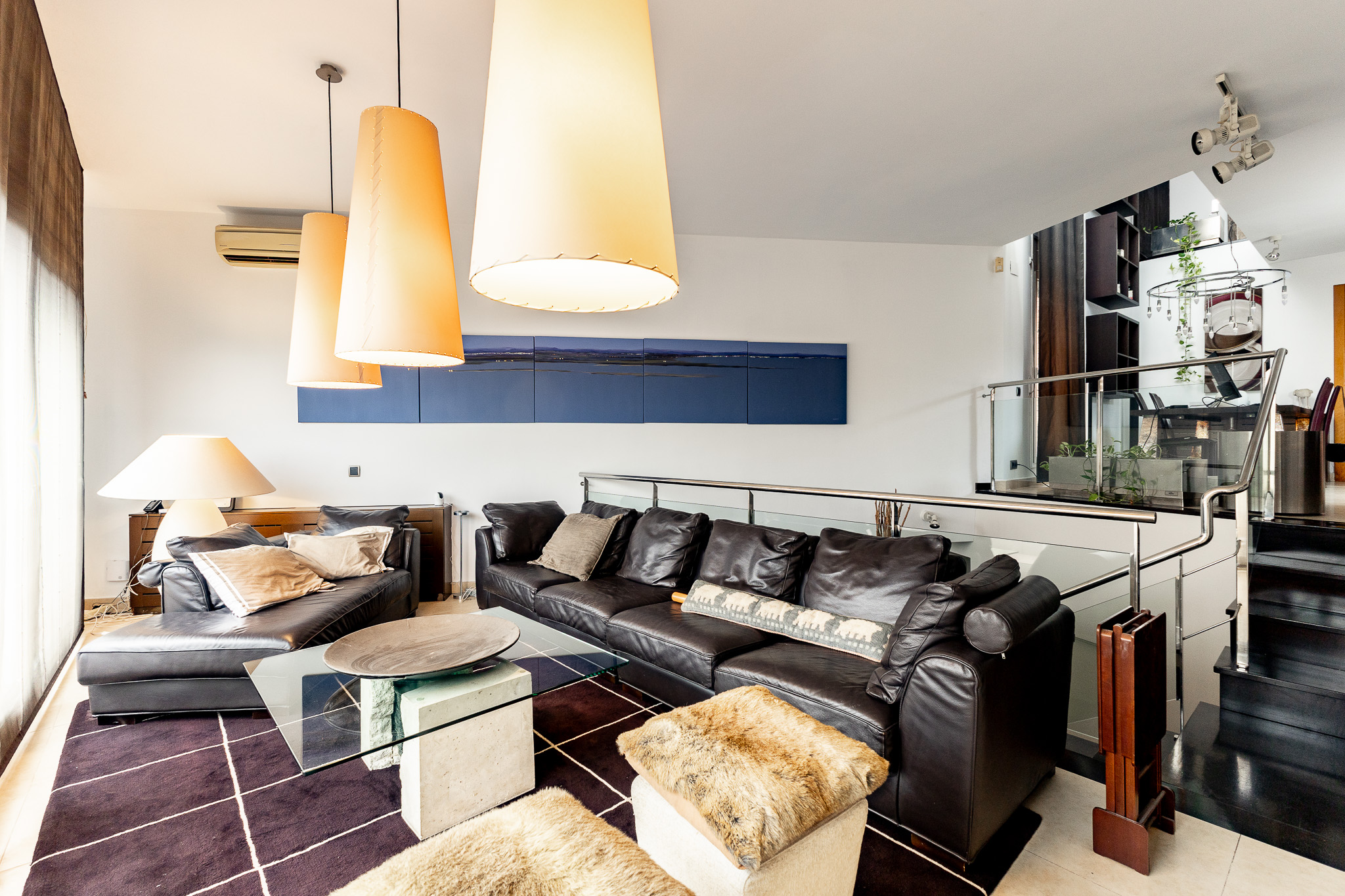 Modern living room with black leather sofas, a glass coffee table, large pendant lights, and a dark grid-pattern rug.
